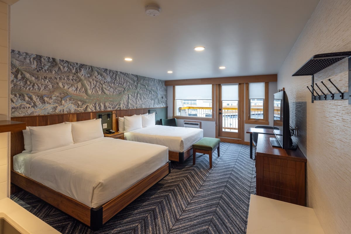 A hotel room with two beds, a desk, a chair, a television, and a large window offering a view of the outside.