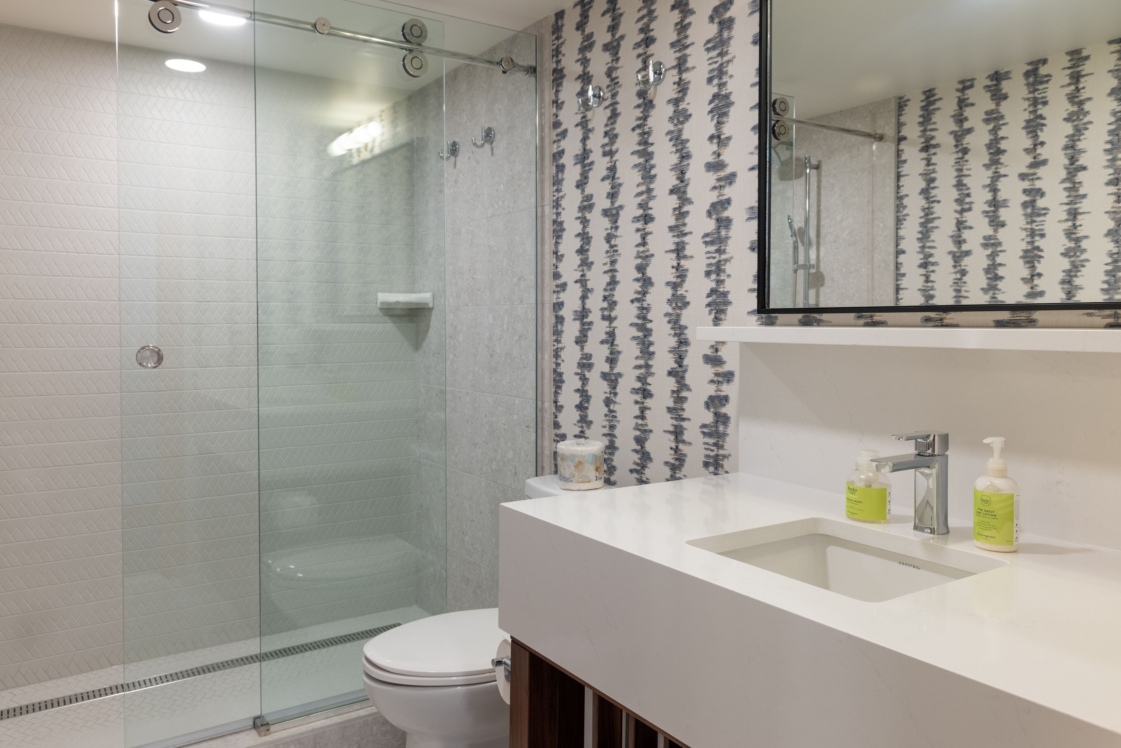 Un bany modern amb una cabina de dutxa de vidre, un lavabo blanc amb dues ampolles de xampú i un mirall gran sobre el lavabo.