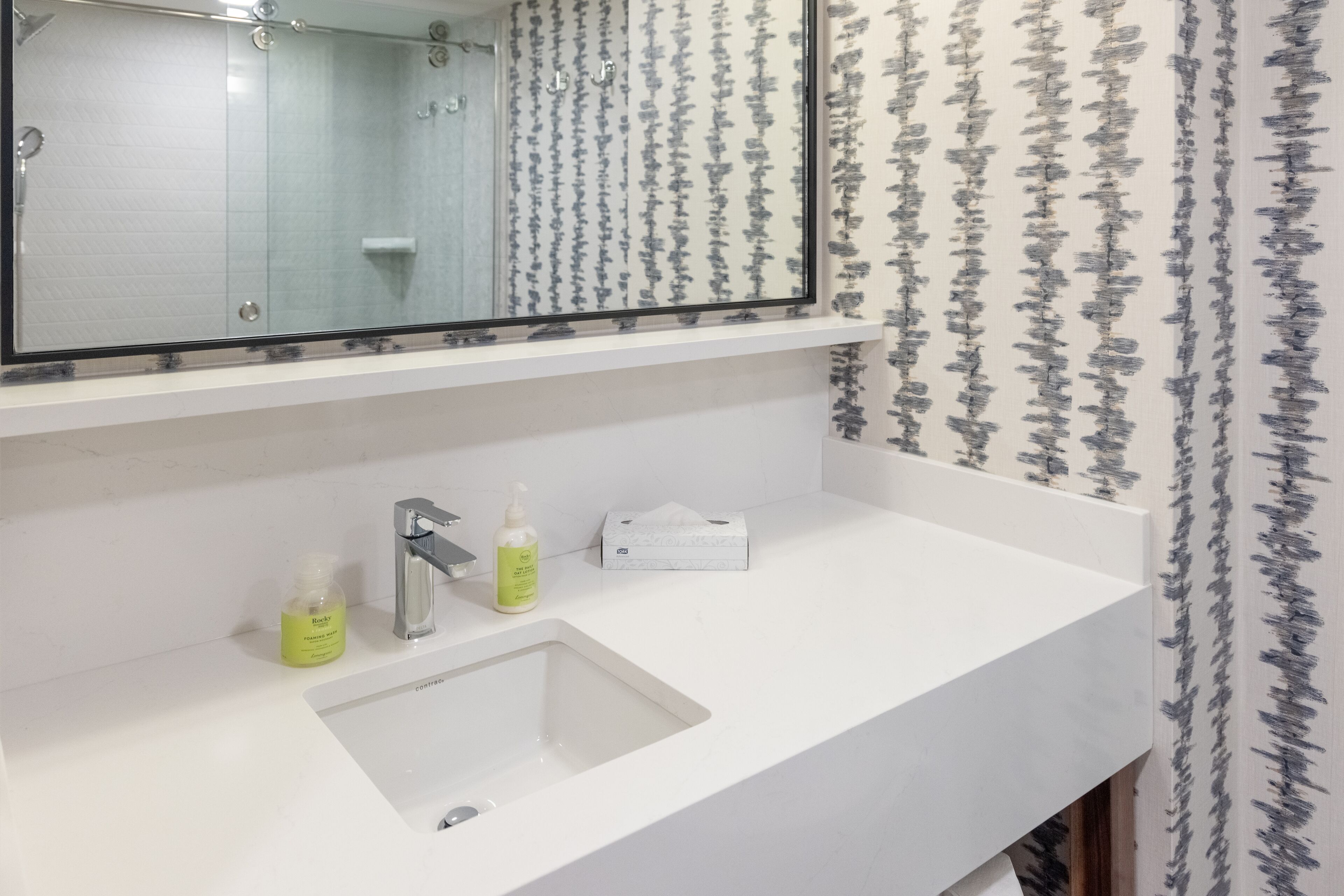 Un baño con un lavamanos blanco, un espejo y una ducha. Hay dos botellas de loción sobre el lavamanos.