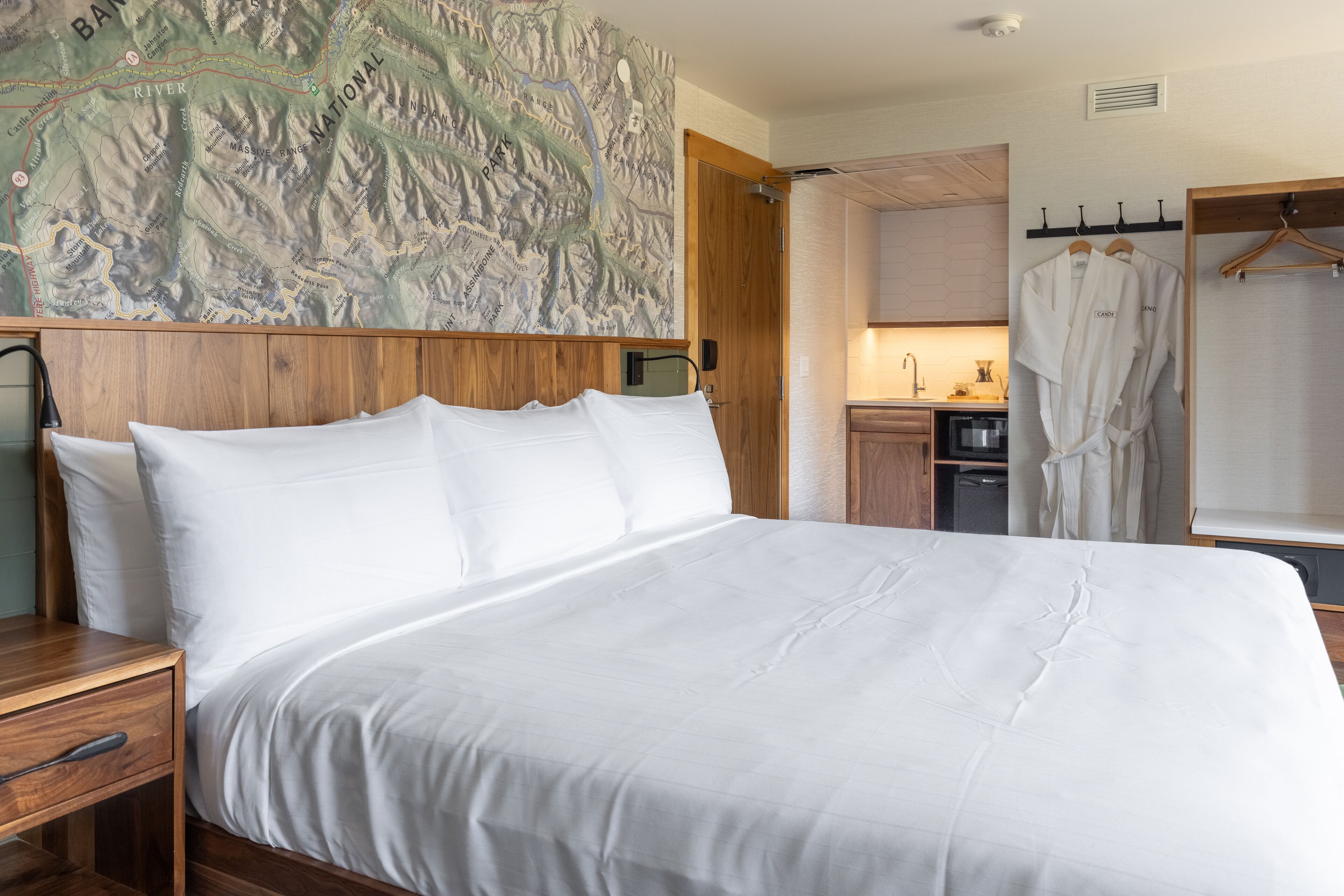 Ein Hotelzimmer mit einem großen Bett, einem Holz-Kopfteil, einer Karte an der Wand, einer kleinen Küchenzeile und einem Bademantelständer.