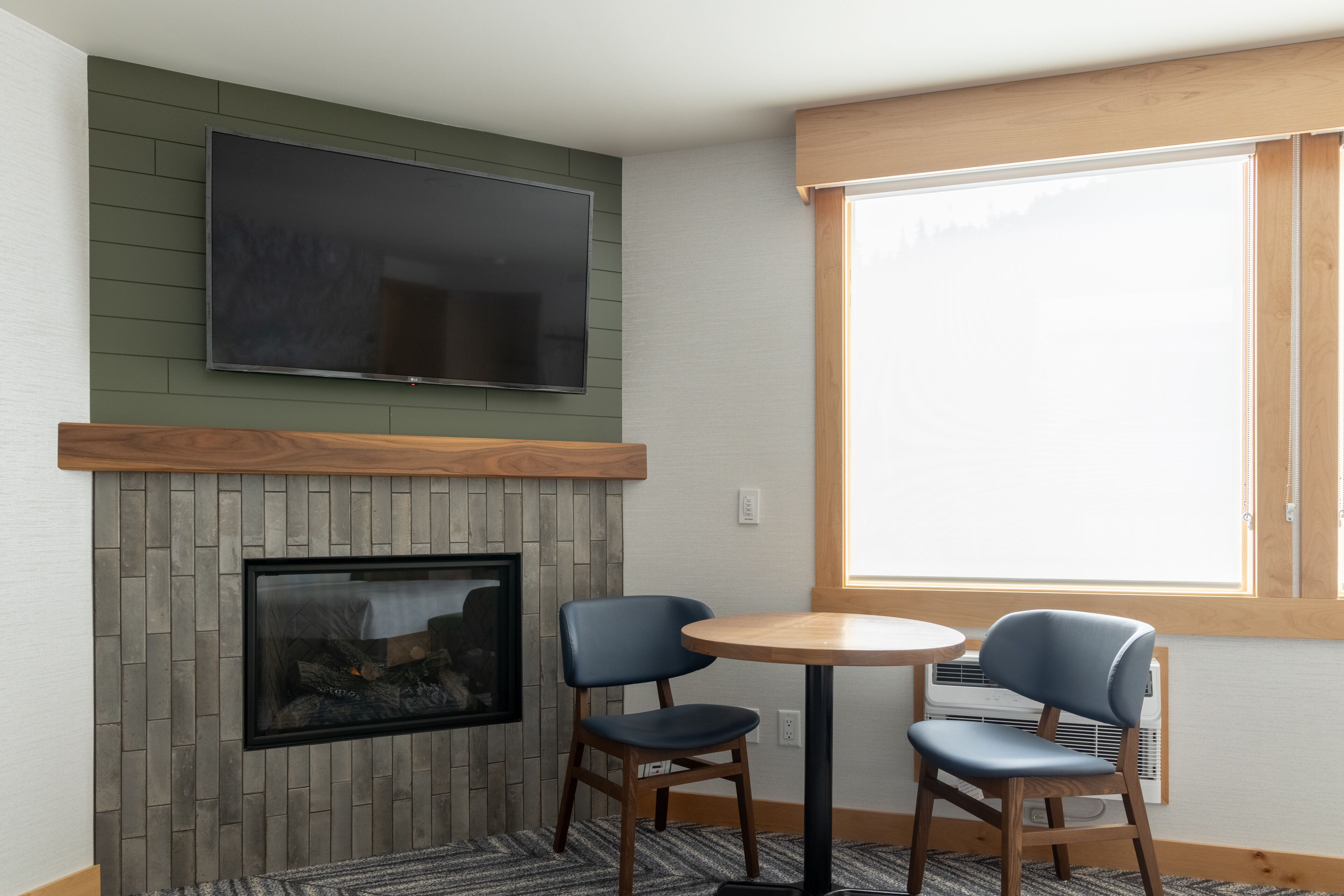 A room with a mounted TV, a fireplace, two chairs, a small table, and a window with a view.