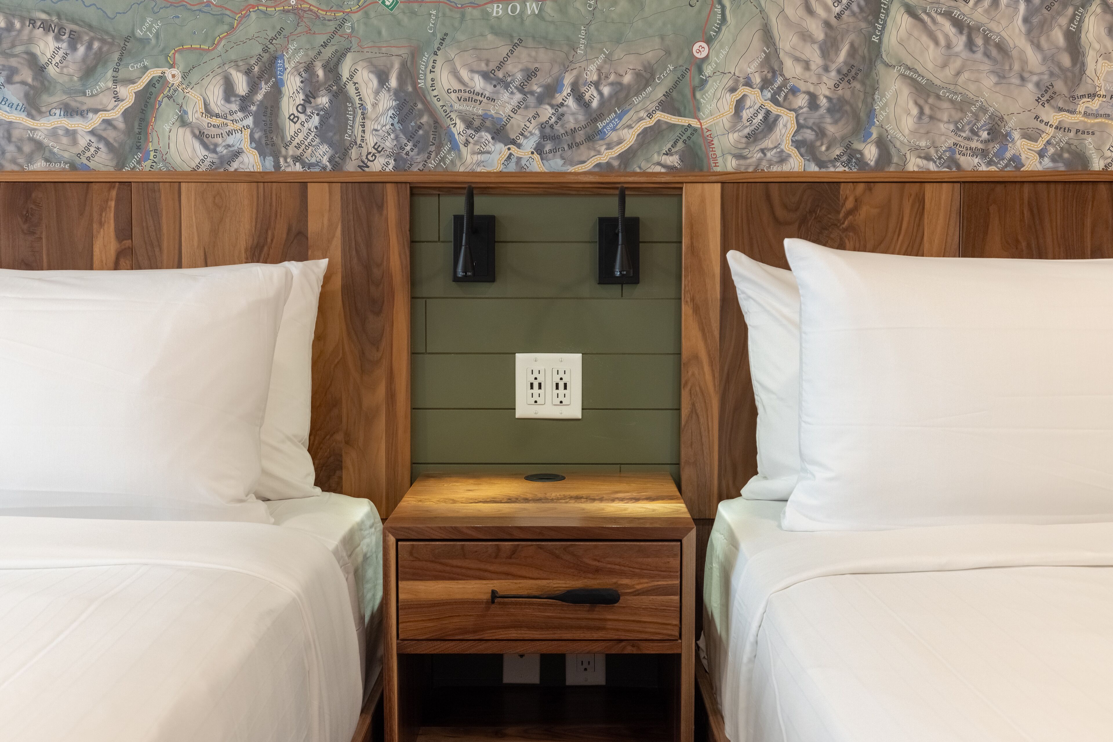 Una habitación con dos camas, un cabecero de madera, un mapa en la pared y una mesita de noche con cajón.