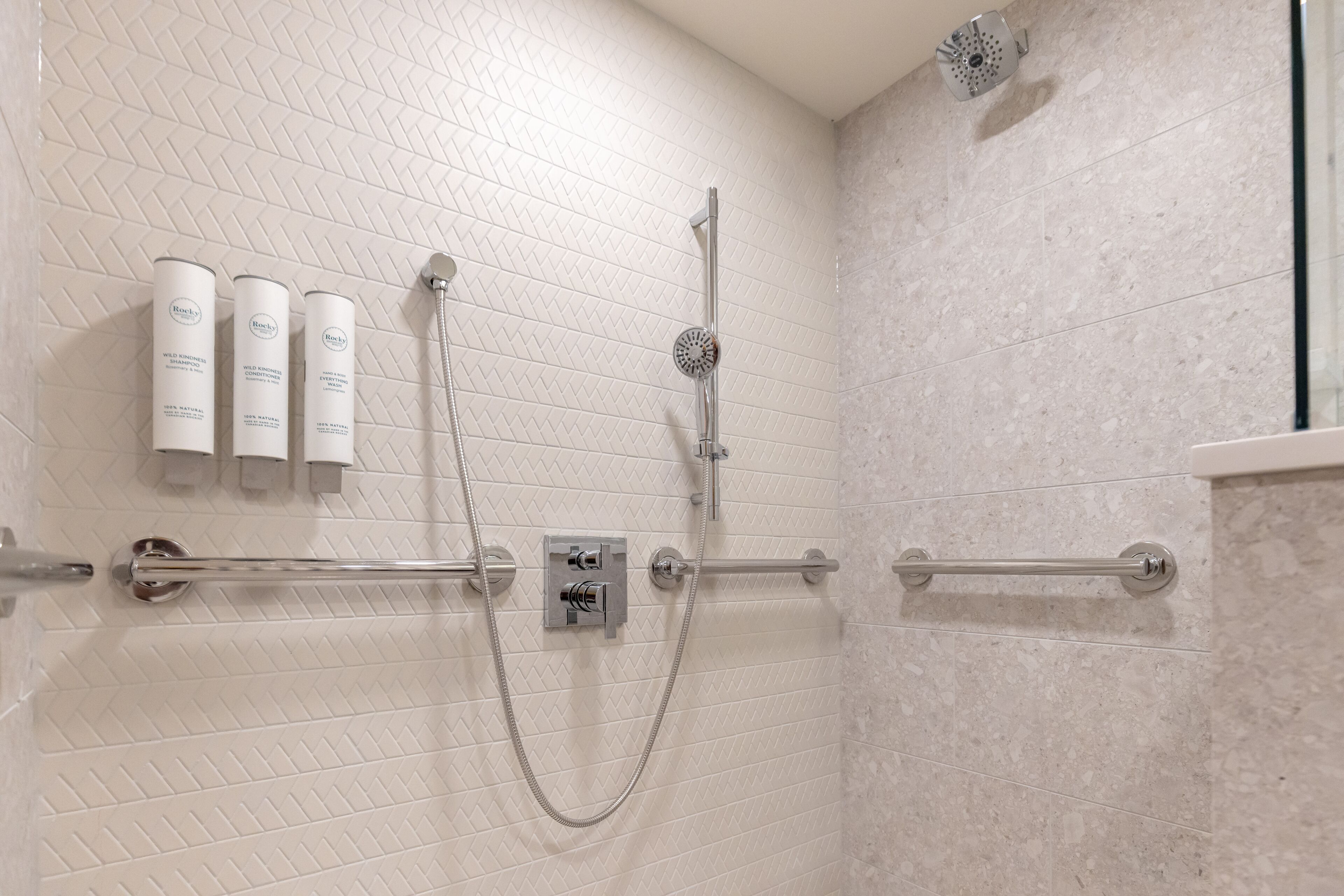 A shower area with a handheld showerhead, a wall-mounted control panel, and three bottles of shampoo.
