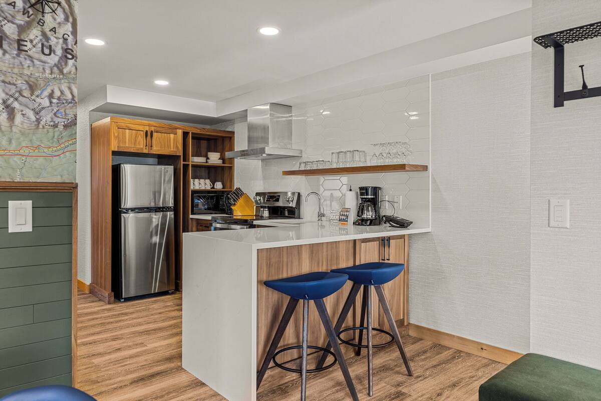 A modern kitchen with a stainless steel refrigerator, wooden cabinets, a white countertop, and blue bar stools.