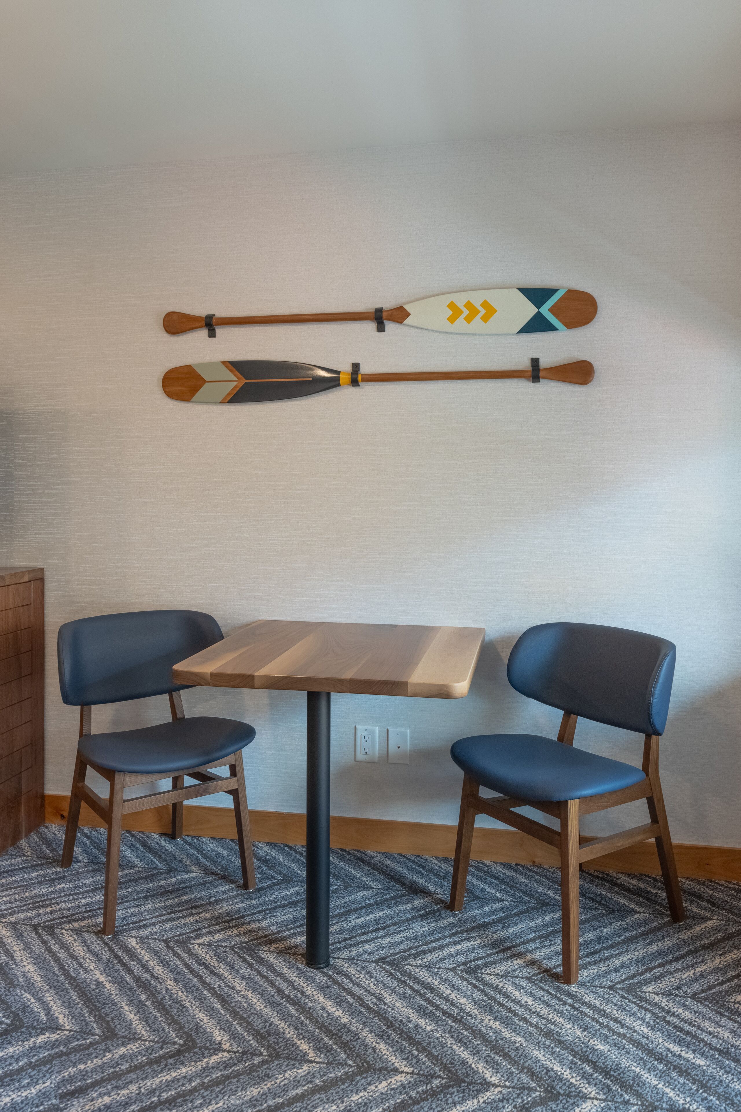 Una habitación pequeña con una mesa de madera y dos sillas azules. Encima de la mesa, hay dos palas decorativas en la pared.