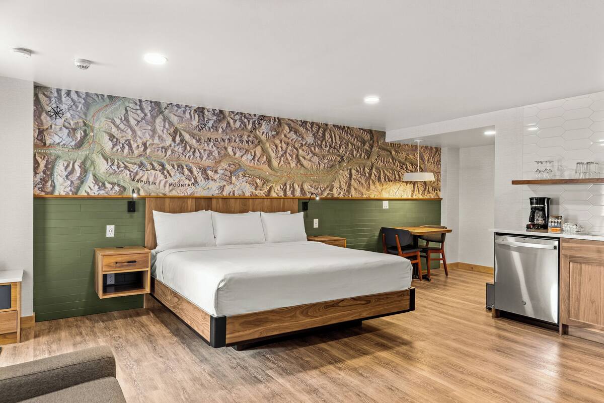 A modern bedroom with a wooden bed, a green accent wall, a map mural, a small desk, and a built-in dishwasher.