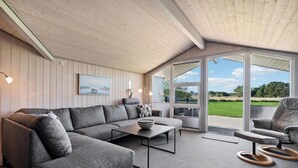 Una sala de estar moderna con un sofá seccional gris, una mesa de centro y una mesa de comedor. Hay un amplio ventanal corredero que da a un área exterior con vistas a un campo verde y árboles.