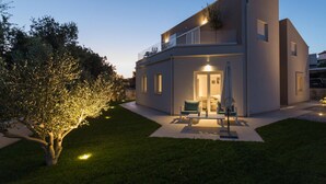A modern two-story house with a well-lit entrance, a patio with outdoor furniture, and a tree illuminated by lights. - Beautiful home in Barbariga (Barbariga)