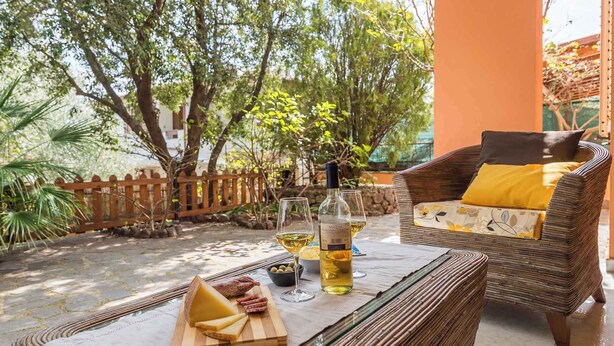 A wicker chair with a yellow cushion, a glass table with a bottle of wine, and a wooden board with cheese and olives. - Villa in a quiet location (Cala Gonone)