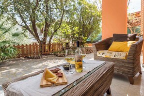 Una silla de mimbre con un cojín amarillo, una mesa de vidrio con una botella de vino y una tabla de madera con queso y aceitunas.