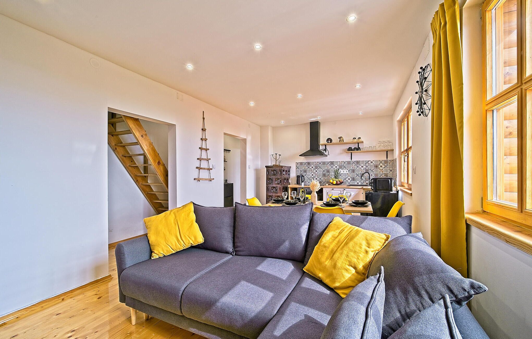 Un salon moderne avec un canapé gris et des coussins jaunes, un escalier et une cuisine en arrière-plan.