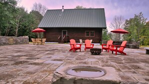 Una cabaña de madera con un patio de piedra, mobiliario exterior y un pozo de piedra.