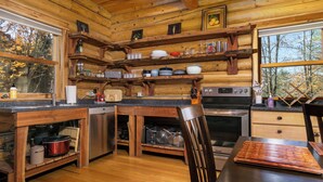 Cocina rústica de cabaña con paredes de madera, una mesa de comedor de madera, un fregadero, una estufa y estantes con platos y utensilios.