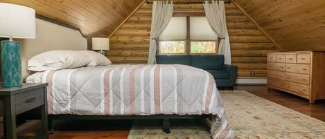A cozy bedroom with a wooden ceiling, a bed with patterned bedding, a nightstand with a lamp, a dresser, and a window with curtains.