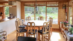 Interior de madera con una mesa redonda rodeada de sillas, una araña y ventanas amplias que dan a una zona boscosa.