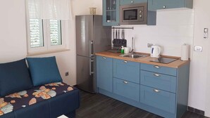 Cocina compacta con refrigerador, microondas e instalación de lavamanos. Hay una ventana con persianas y un sofá con un cojín estampado floral.
