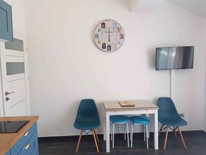 A room with a white wall, a round clock, a flat-screen TV, a wooden cabinet, a table, and two blue chairs.