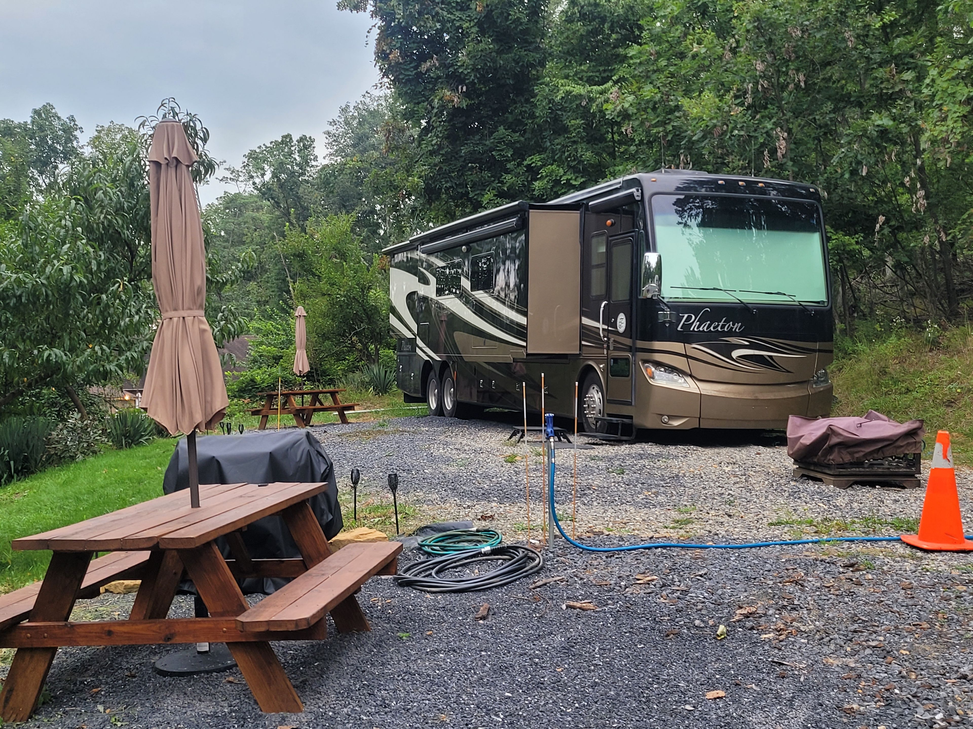 Veículo recreativo (RV) estacionado em um camping com mesas de piquenique e um guarda-sol coberto.