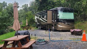 Veículo recreativo (RV) estacionado em um camping com mesas de piquenique e um guarda-sol coberto.