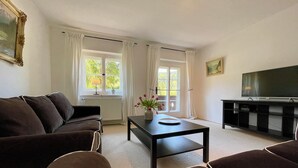 A living room with a sofa, a coffee table with a vase of flowers, a television, and a window with curtains. - Forsthaus Fewo 10 OG (Reit im Winkl)