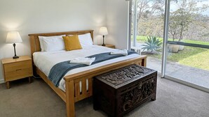 A bedroom with a wooden bed, two bedside tables with lamps, a large window with a view of greenery, and a decorative wooden bench.