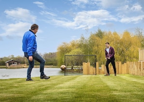 Two people playing soccer on a grassy field with a goalpost, a body of water, and trees in the background.