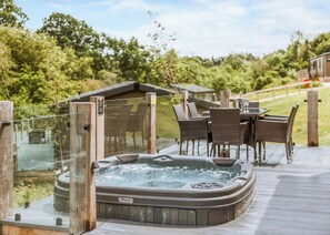 A hot tub on a wooden deck with wicker chairs and a table, surrounded by greenery.