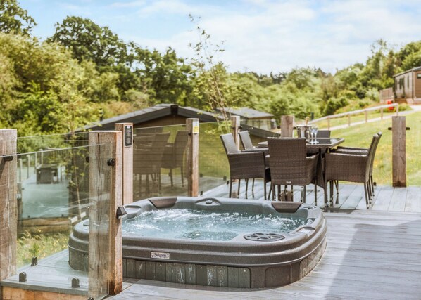 A hot tub on a wooden deck with wicker chairs and a table, surrounded by greenery.