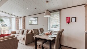 A modern room with a dining table, chairs, a sofa, and a large window with blinds. - Sycamore Lodge VIP Hot Tub (Bridgnorth)