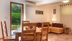 A living room with a wooden dining table, chairs, a sofa, and a lamp. There is a door leading to a garden, and an air conditioning unit on the wall. - Holiday apartment with a beautiful garden for shared use (Vinkuran)