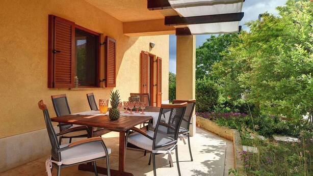 An outdoor patio with a wooden table and chairs, surrounded by a yellow wall and wooden shutters. - Holiday apartment with a beautiful garden for shared use (Vinkuran)
