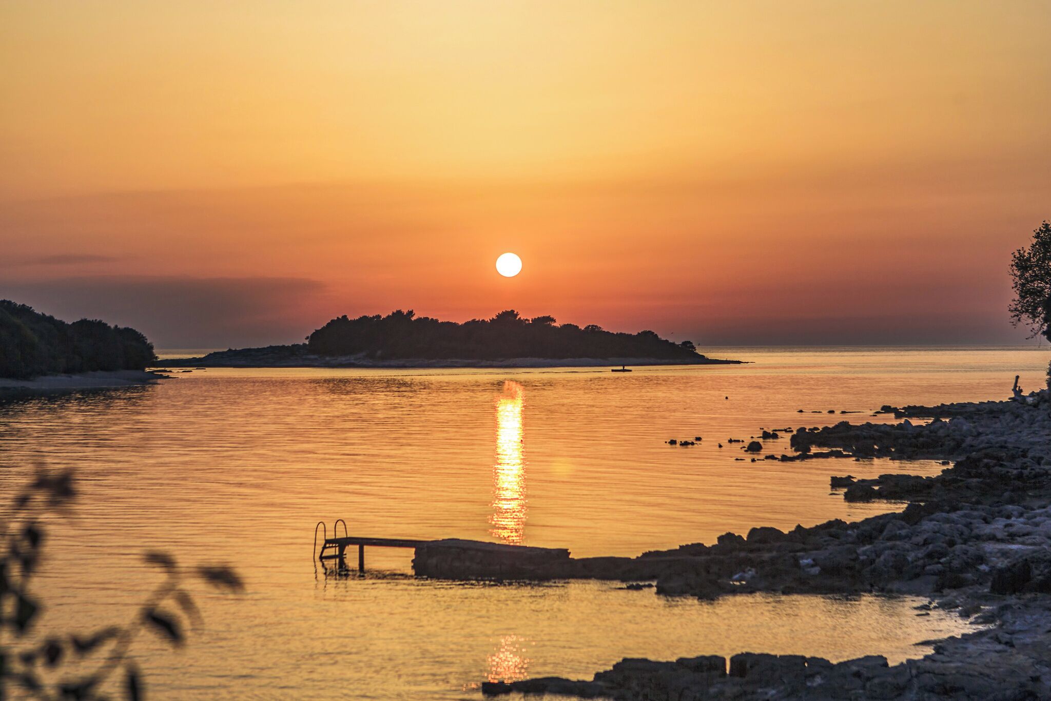 Ein Sonnenuntergang über ruhigem Wasser mit einer felsigen Küste und einer kleinen Insel in der Ferne.
