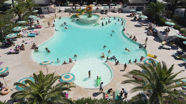 Uma grande piscina ao ar livre rodeada por espreguiçadeiras e guarda-sóis, com um escorrega e um edifício com telhado de palha ao fundo.