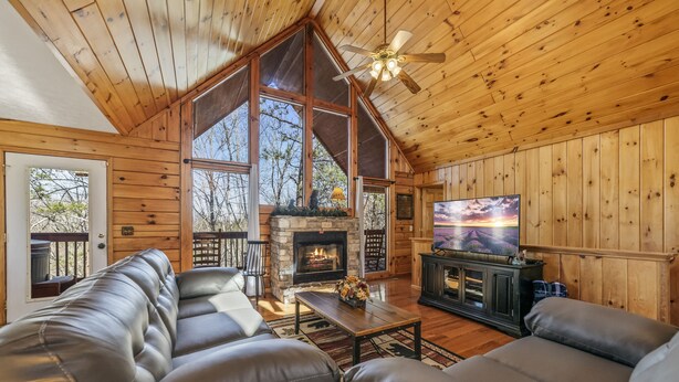 Interior de una cabaña de madera con chimenea, sofás de cuero, televisor y un ventanal con vistas al exterior.