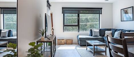 A modern living room with a sofa, a coffee table, and a dining table. There are large windows with a view of trees outside.