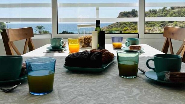 A table with breakfast items and a bottle of water, set against a large window with a view of the sea and hills.