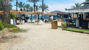 A beachside restaurant with outdoor seating, palm trees, and a clear sky.