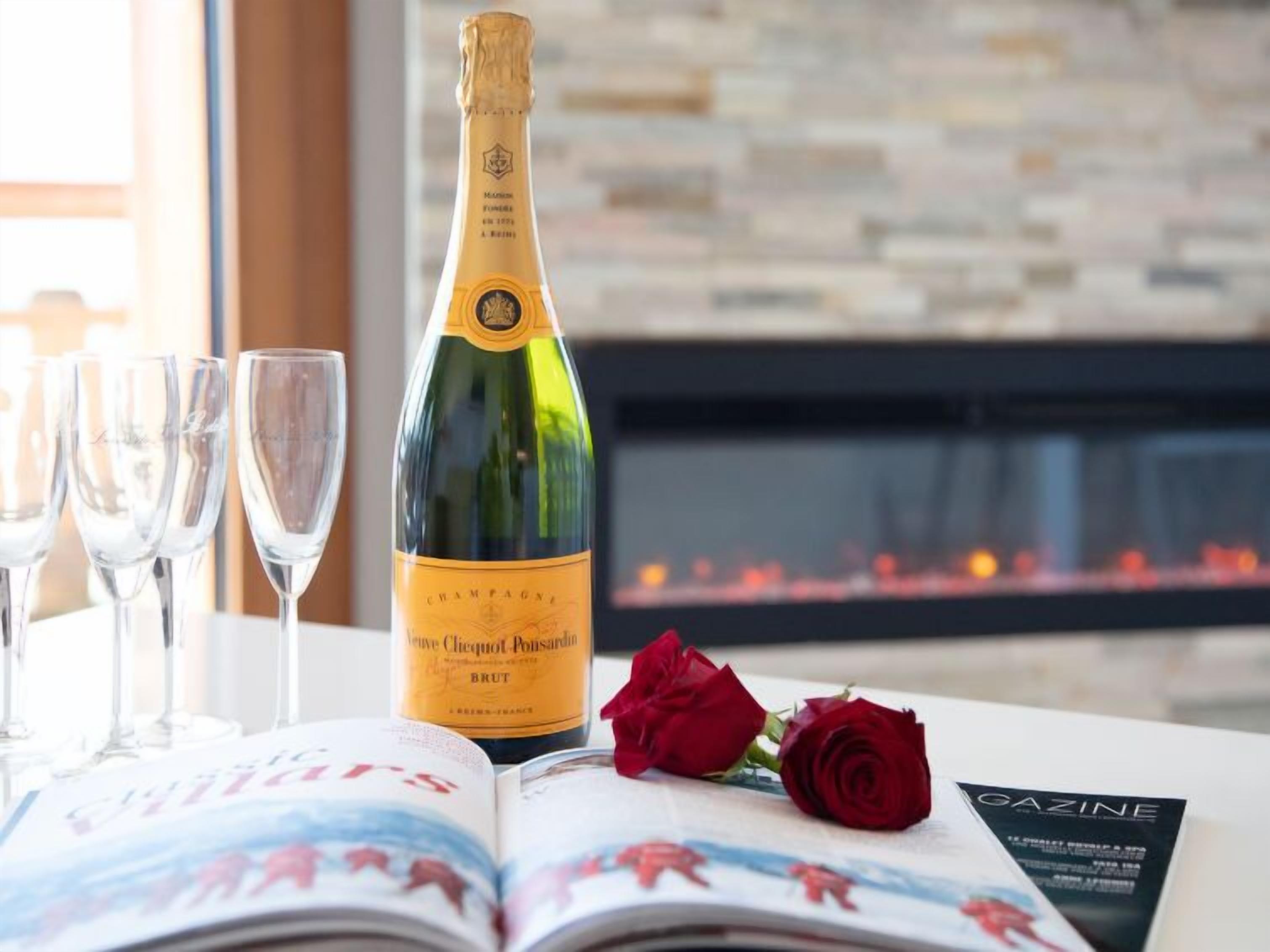 A bottle of champagne, four glasses, a red rose, and a magazine on a table with a fireplace in the background.
