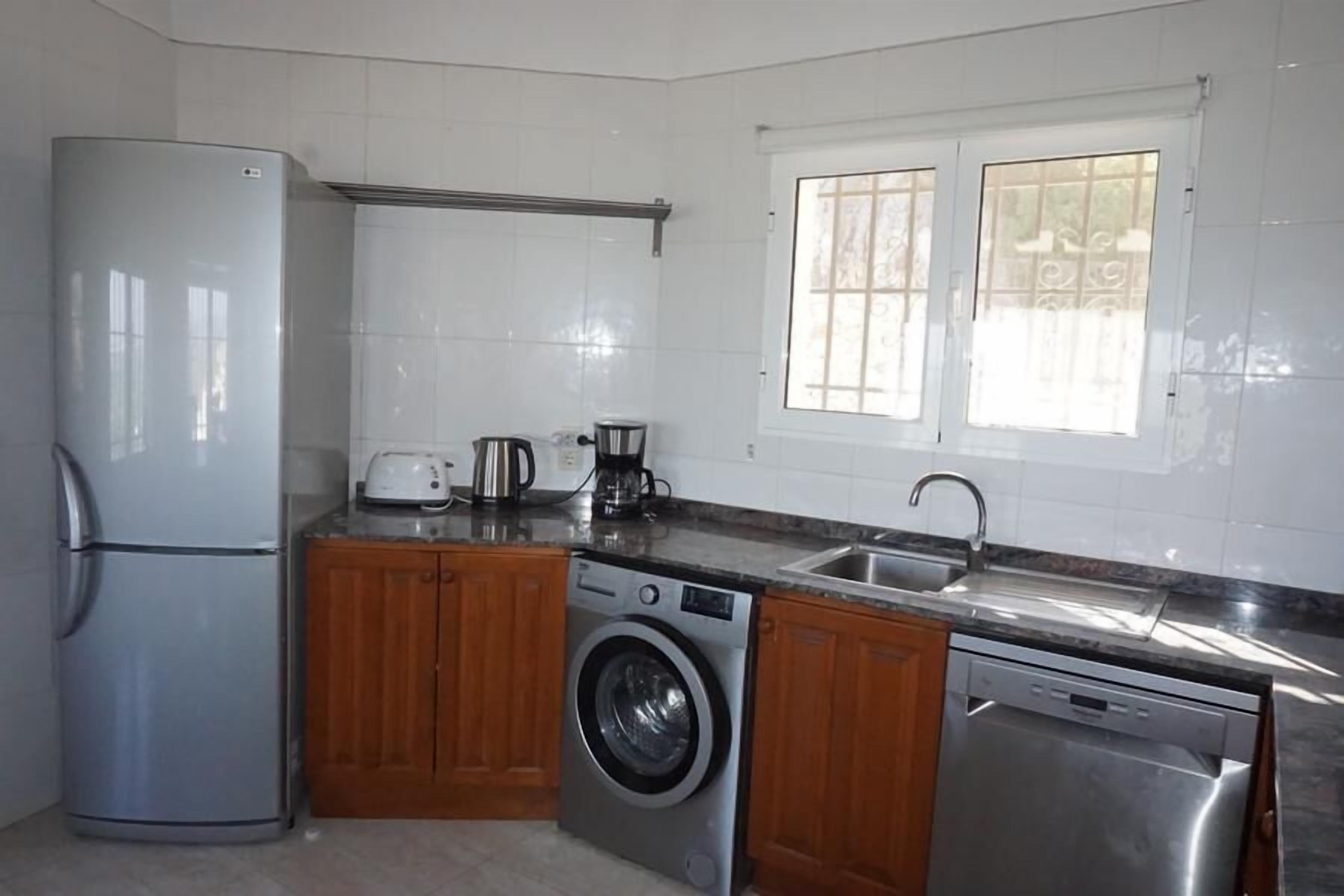 A compact kitchen with a refrigerator, washing machine, dishwasher, and a window with blinds.