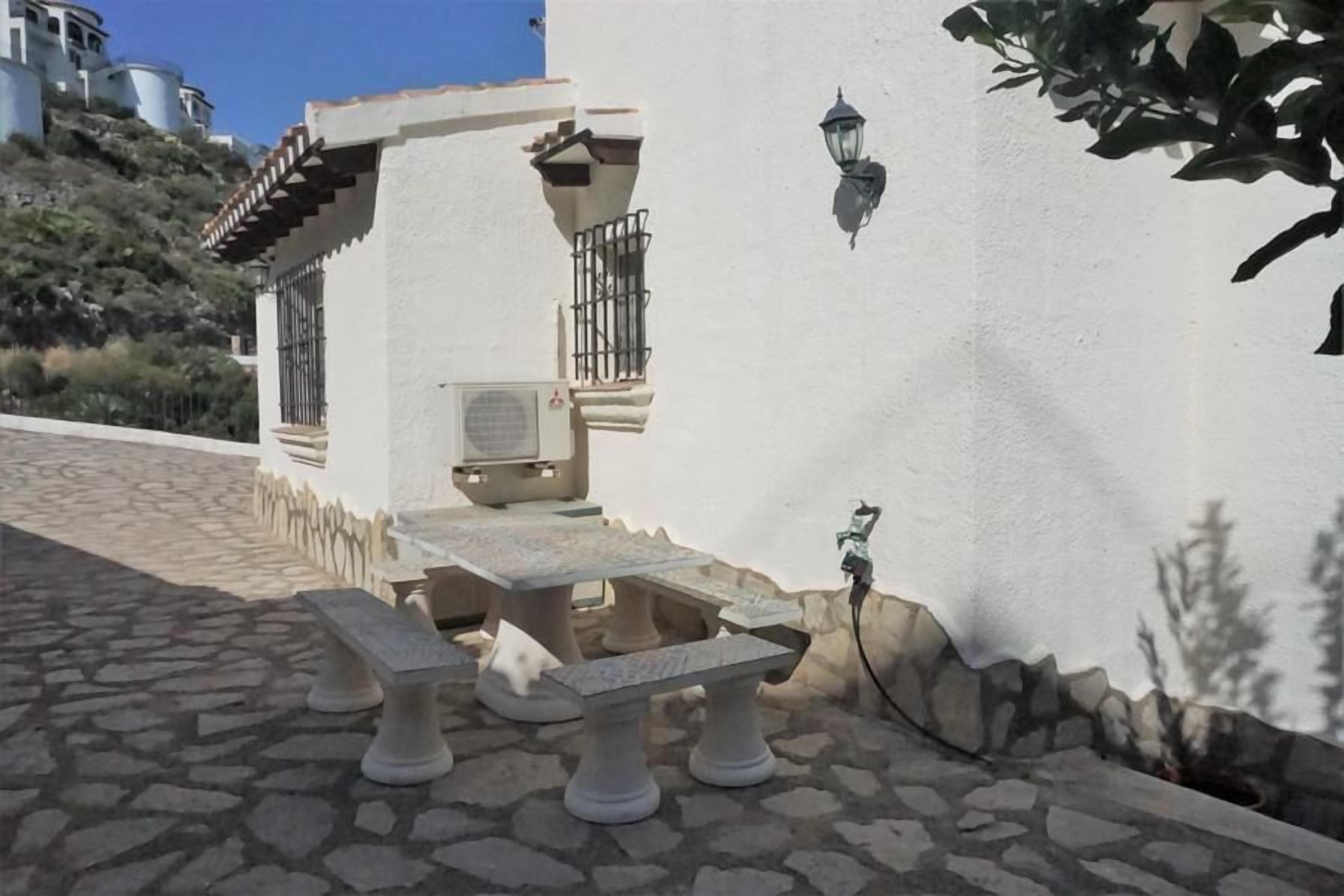 A stone patio with a table and benches, a wall-mounted air conditioning unit, and a wall-mounted lamp.