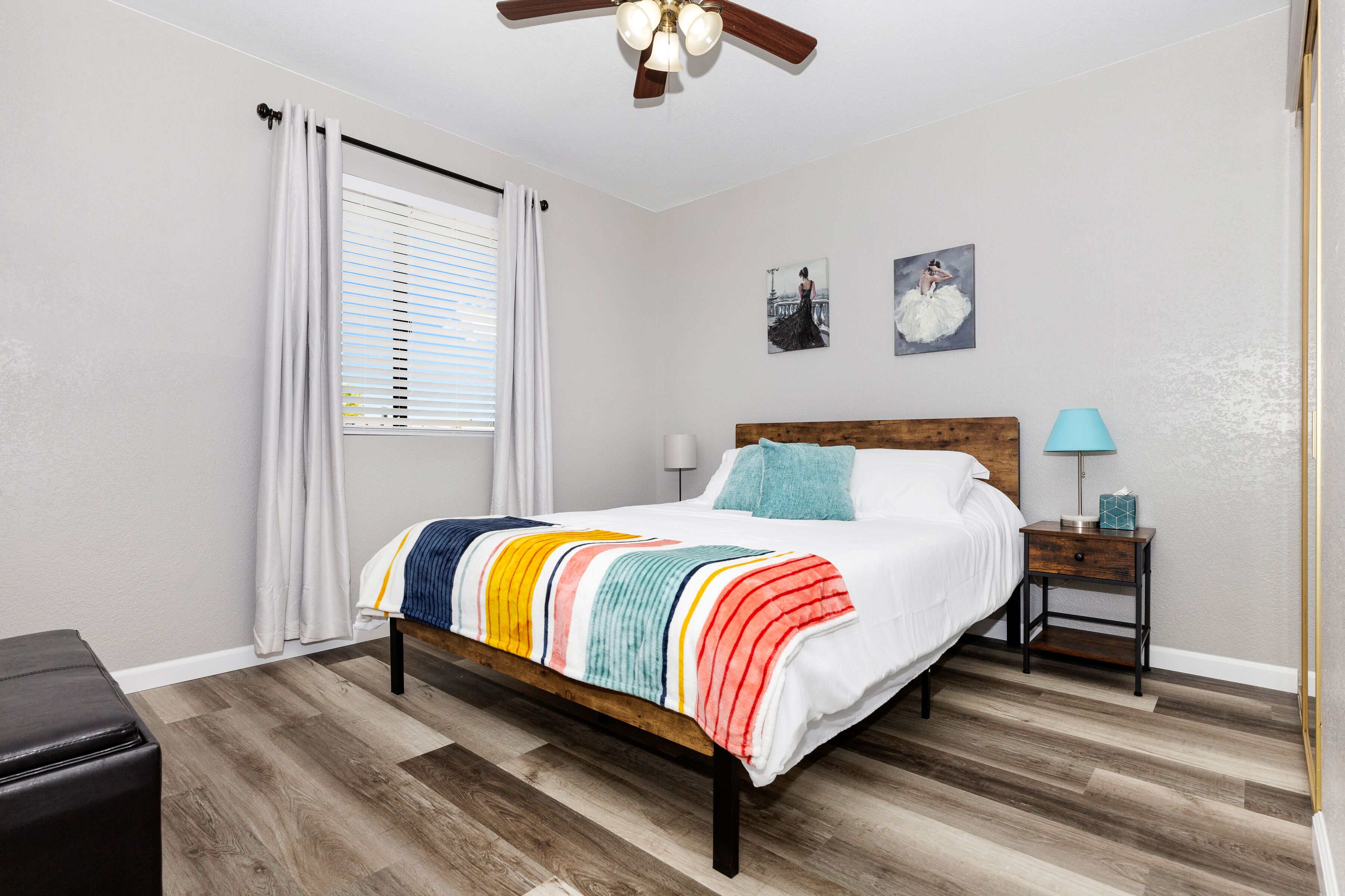 Un dormitorio con una cama de madera, ropa de cama blanca, una manta a rayas coloridas, una mesita de noche con una lámpara y dos cuadros en la pared.