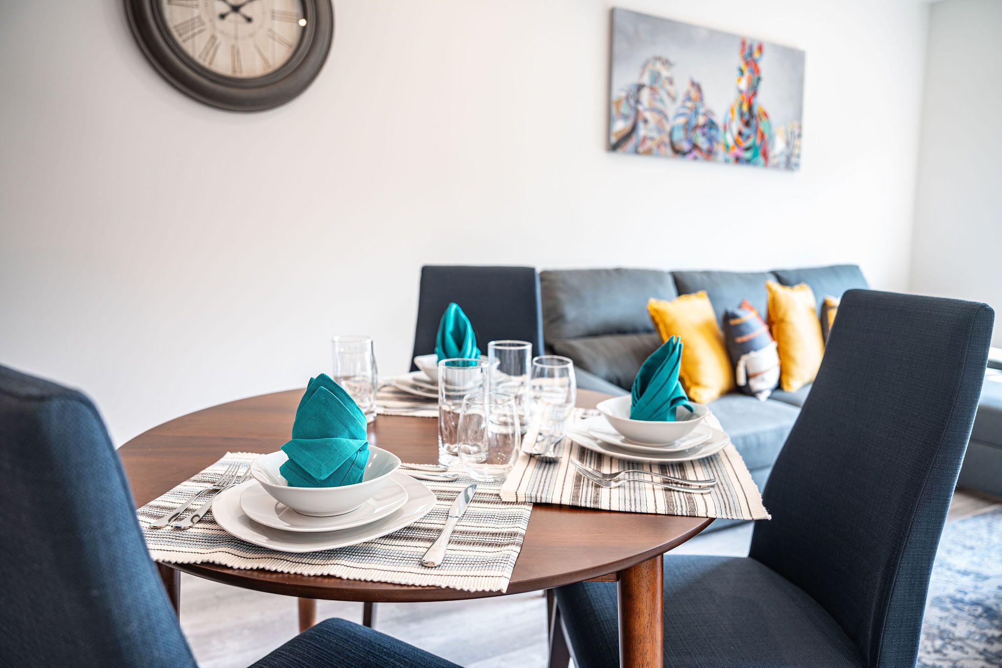 Une table à manger dressée avec des assiettes, des verres et des couverts, une horloge au mur et un canapé avec des coussins à l’arrière-plan.