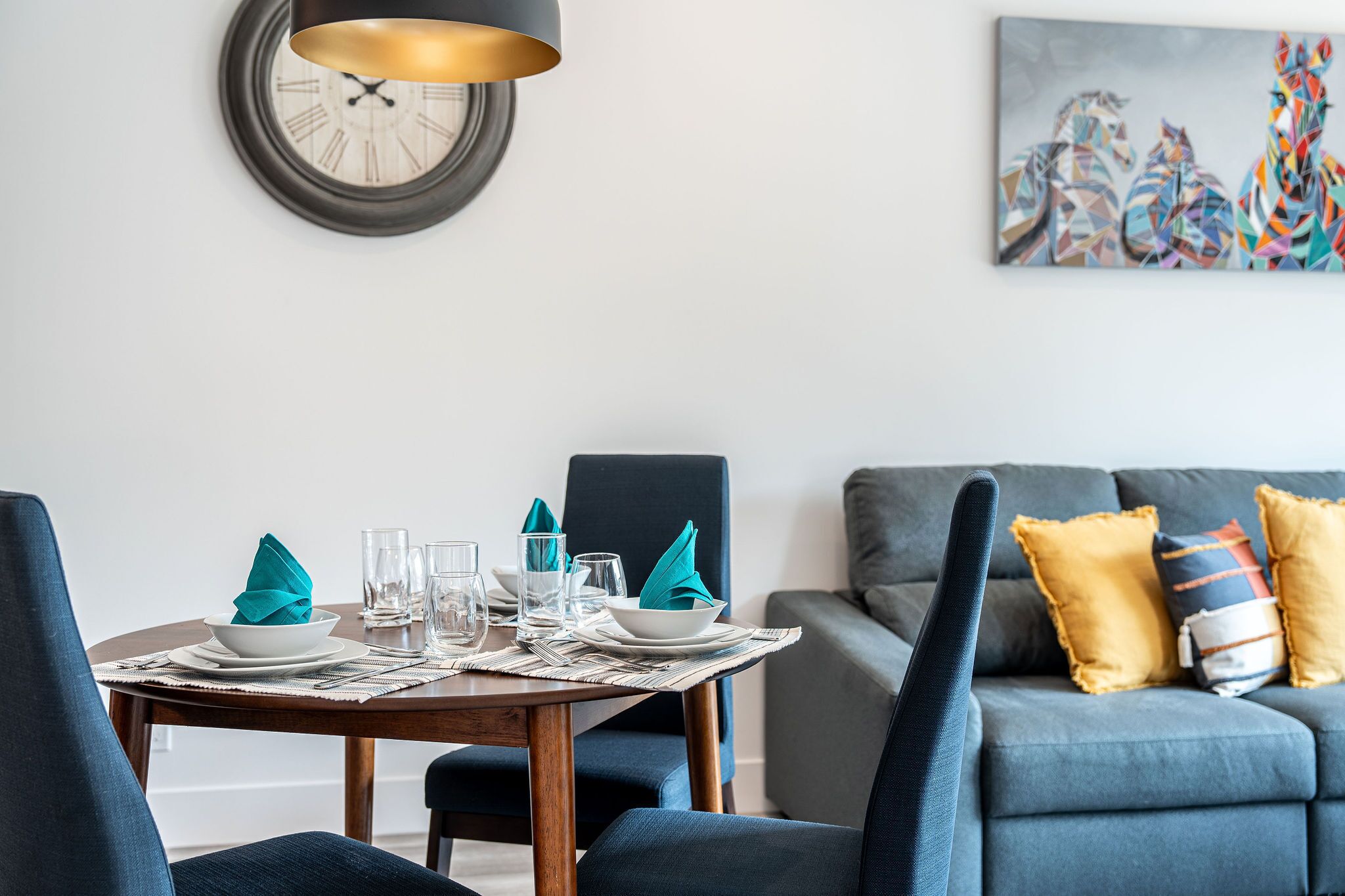 A dining area with a round table set for four, a modern clock on the wall, and a colorful abstract painting above the sofa.