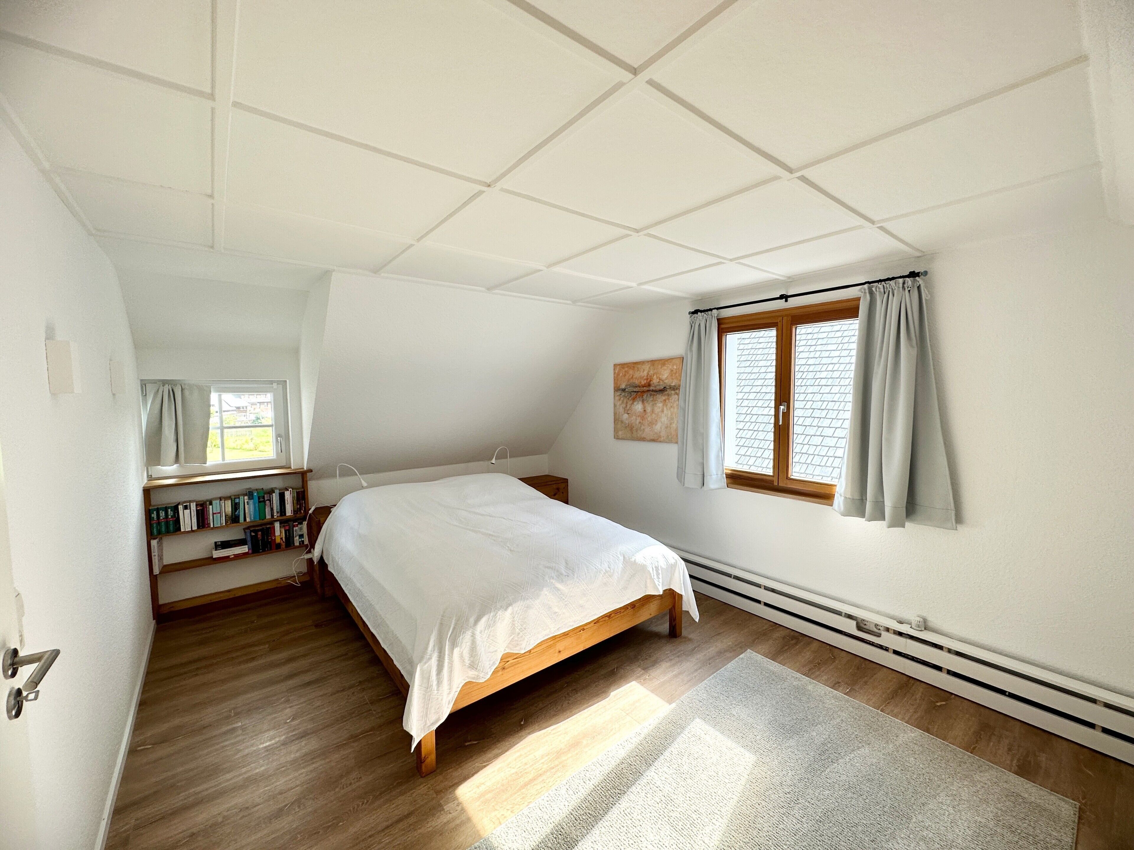 Un dormitorio con cama, ventana con cortinas, radiador y estantería.