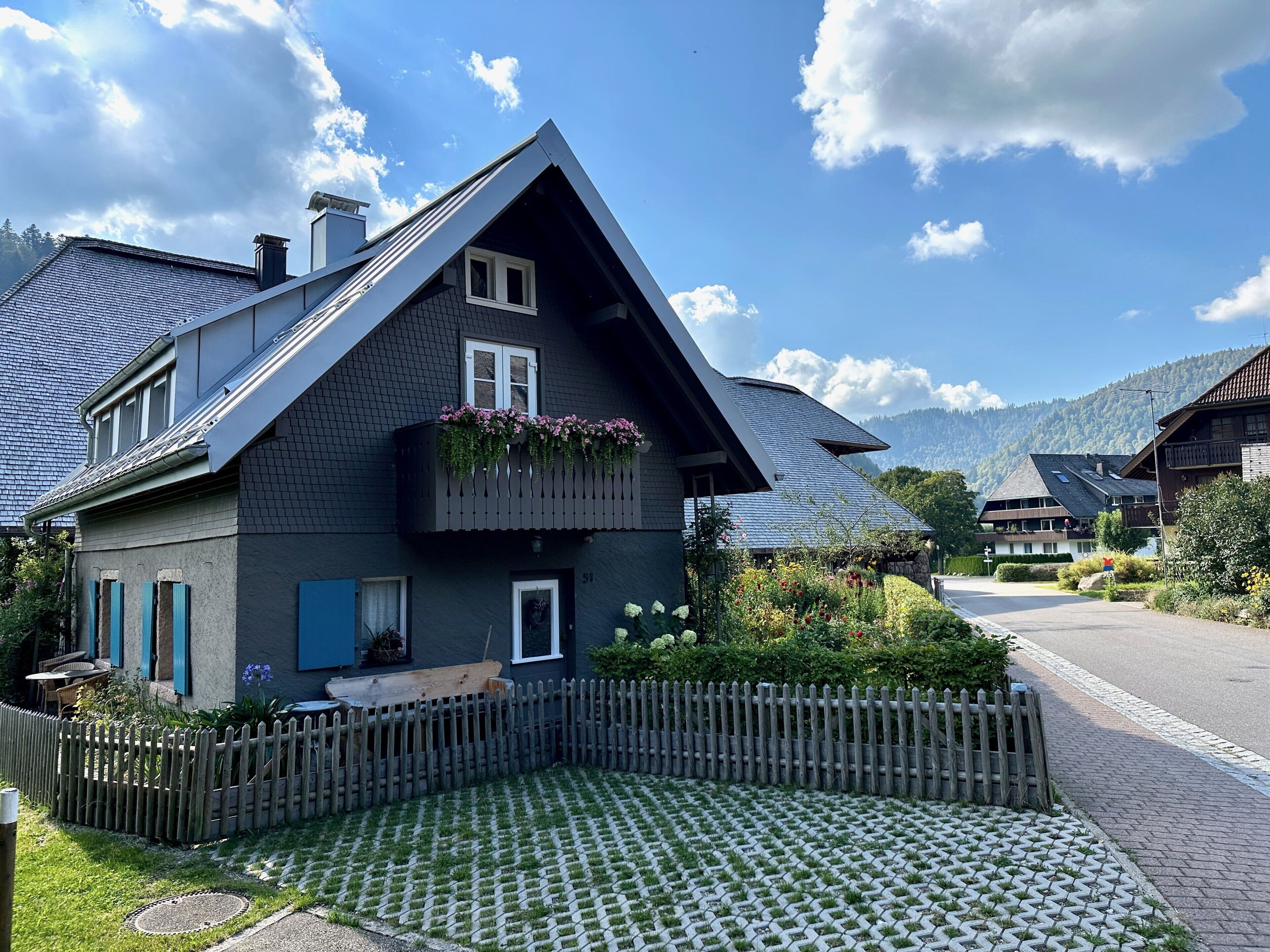 Una casa con techo inclinado, un balcón lleno de flores, una valla de madera y un jardín pavimentado.