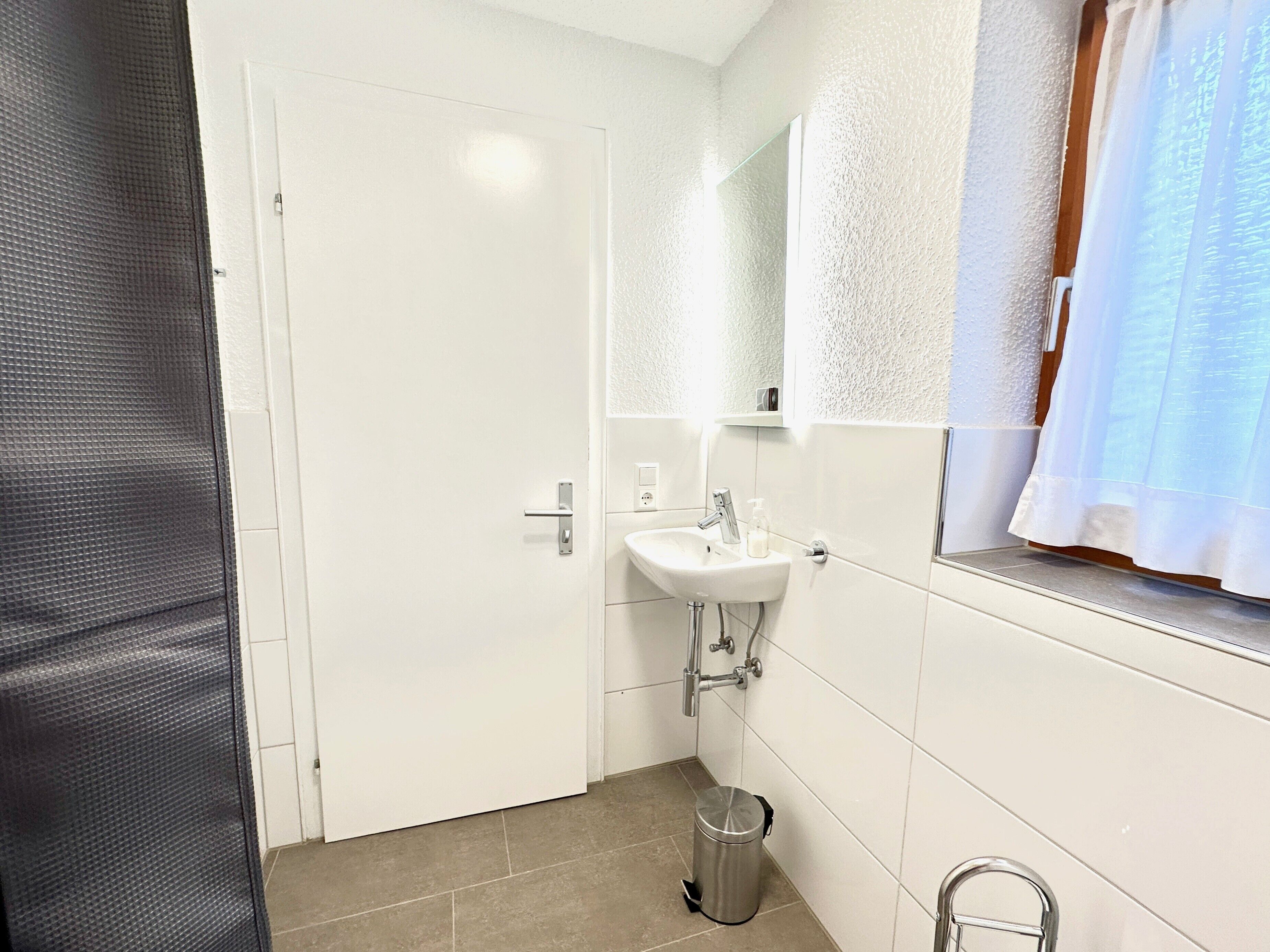 Een badkamer met een wit wastafel, spiegel en een raam met een witte gordijn.