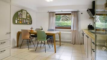 A dining area with a wooden table and chairs, a bulletin board with photos, a window with curtains, and a kitchen area with cabinets and appliances.
