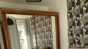 Une chambre avec un grand miroir, un lit recouvert de plusieurs coussins, une table de chevet avec une lampe et un papier peint à motifs.