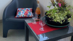 Un balcon avec une chaise noire, un coussin coloré, une bouteille, deux verres et une plante en pot.