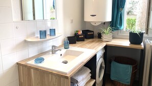 Une salle de bain avec un lavabo, un miroir et une fenêtre avec des rideaux. On trouve des articles de toilette sur le comptoir et une plante sur le rebord de la fenêtre.
